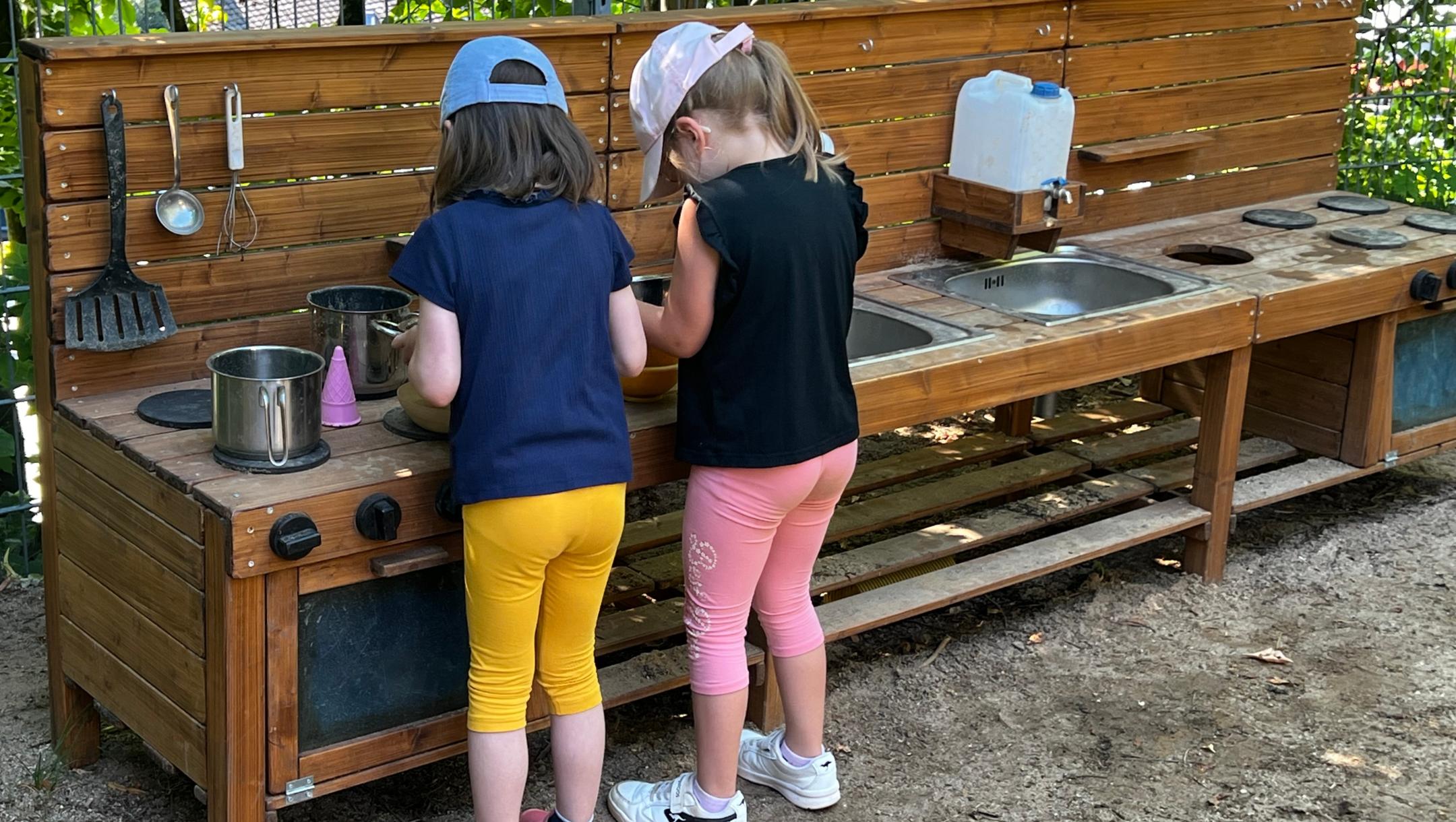 Man sieht zwei Kinder von hinten, die in der Matschküche des Außenbereiches spielen