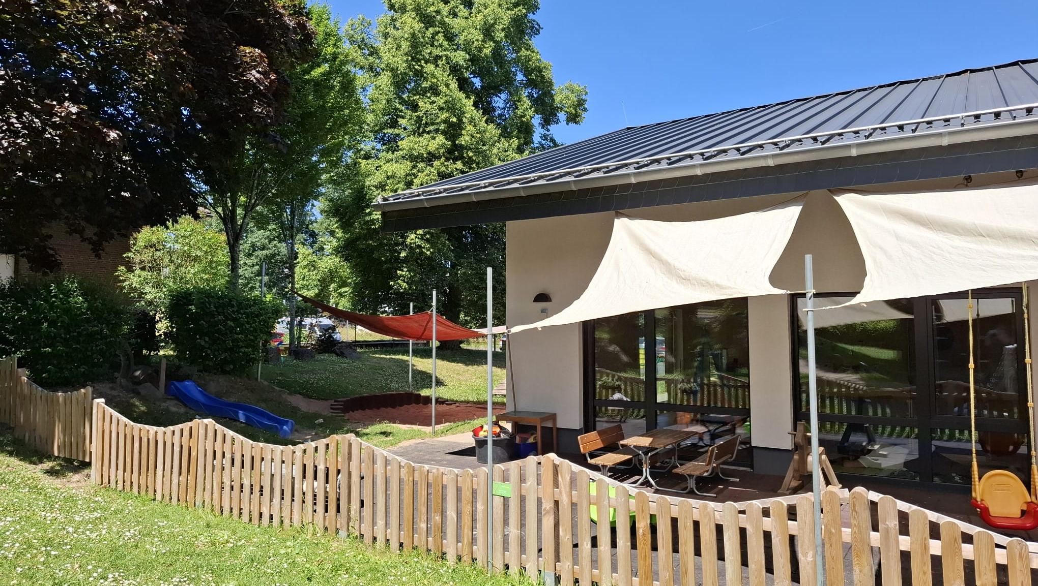 Man sieht das KiTa-Gebäude von Außen mit einem Sonnensegel, unter dem Kinder im Schatten spielen können (c) Katholische KiTa gGmbH Trier Man sieht das KiTa-Gebäude von Außen mit einem Sonnensegel, unter dem Kinder im Schatten spielen können